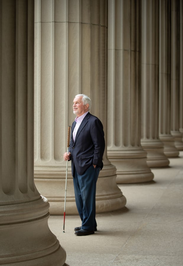 Paul is standing in a blue sport coat among the columns outside of an iconic building on MIT’s campus.  He’s holding his cane and has a confident smile on his face. 